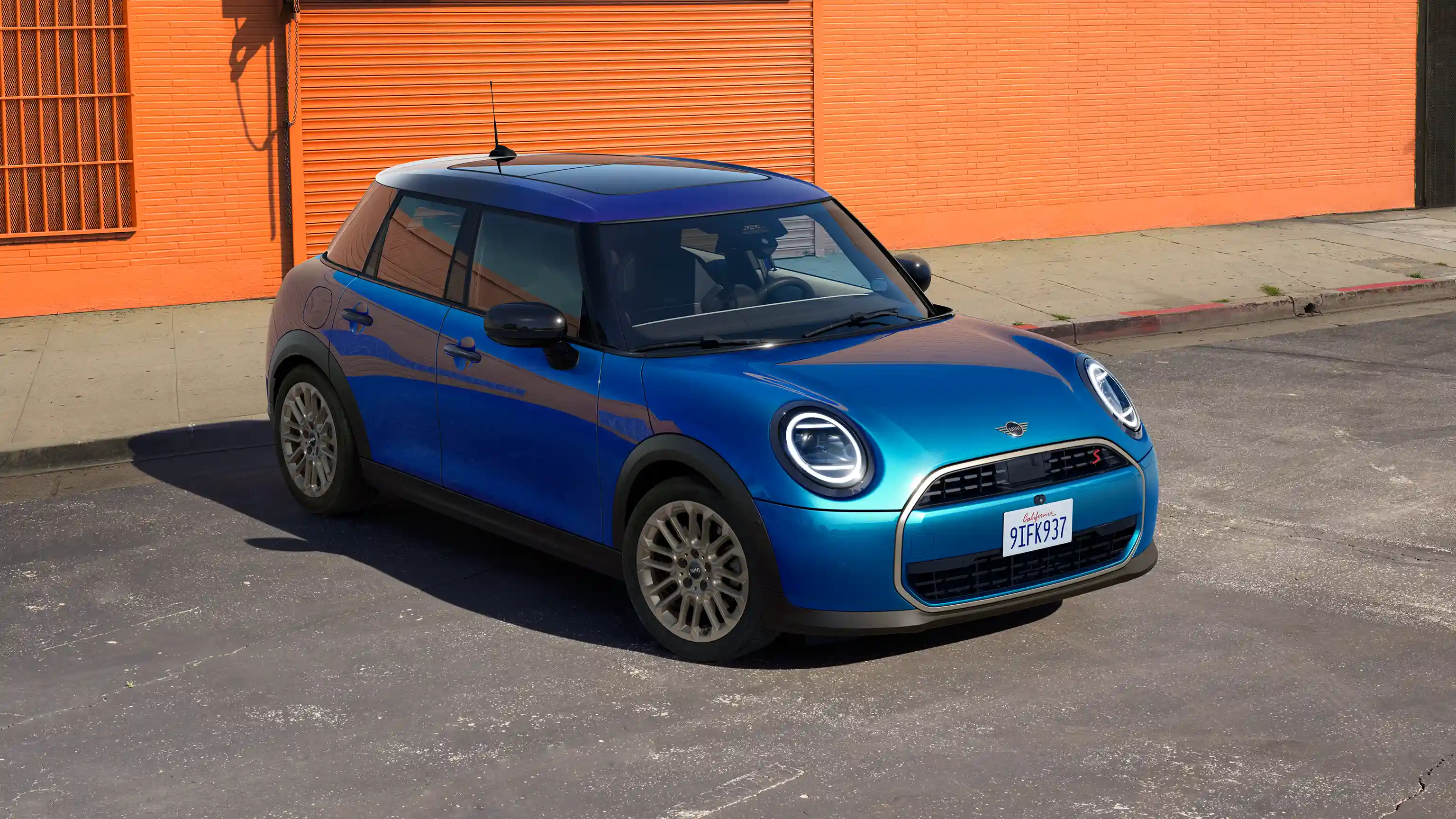 Predný pohľad na MINI Cooper 5-dverové vo farbe Icy Sunshine Blue s viacfarebnou strechou Multitone Blue na ulici pred oranžovou stenou