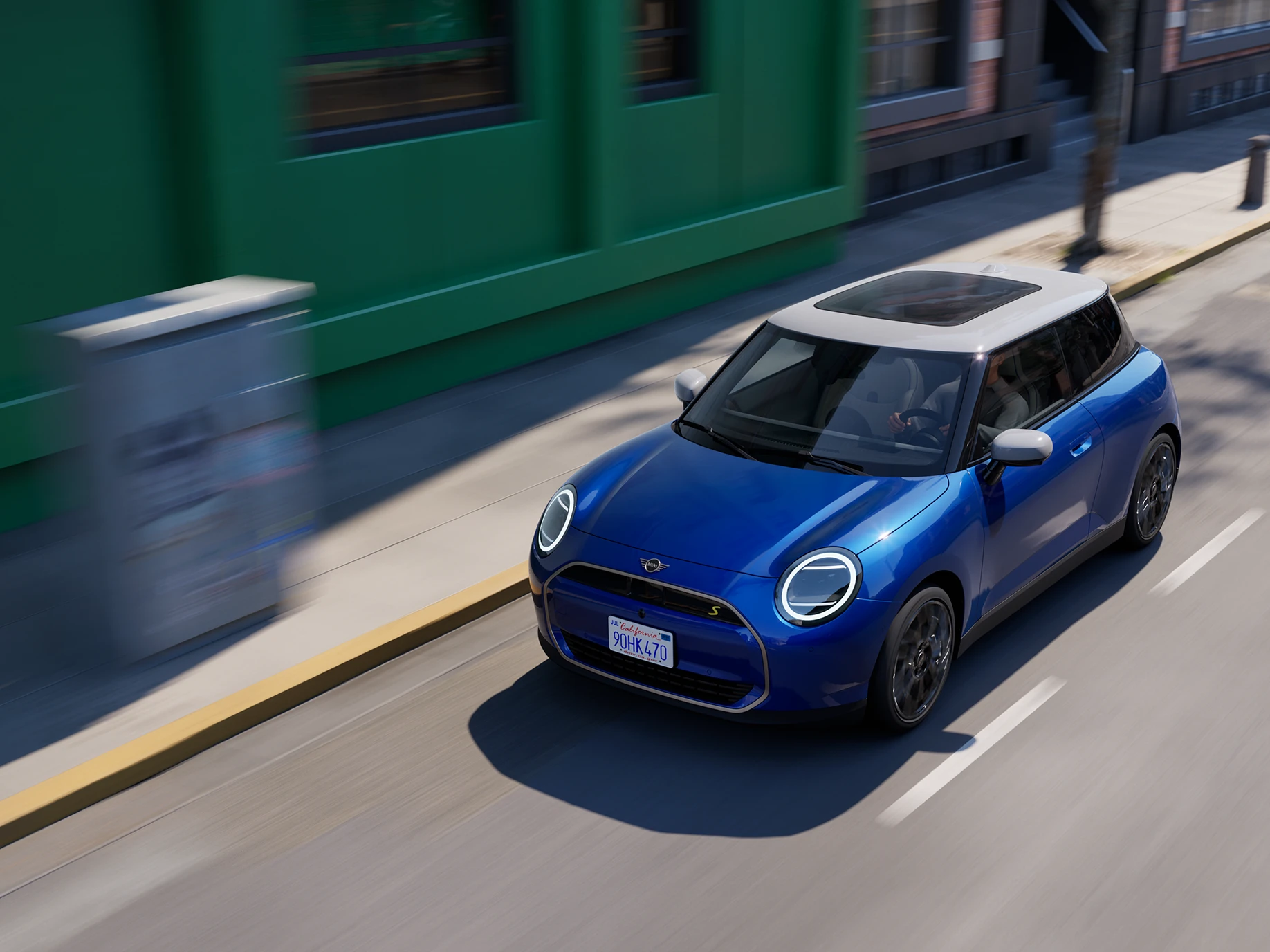 Pohľad zhora na čisto elektrické MINI Cooper vo farbe Blazing Blue so strechou White, počas jazdy na ceste. 