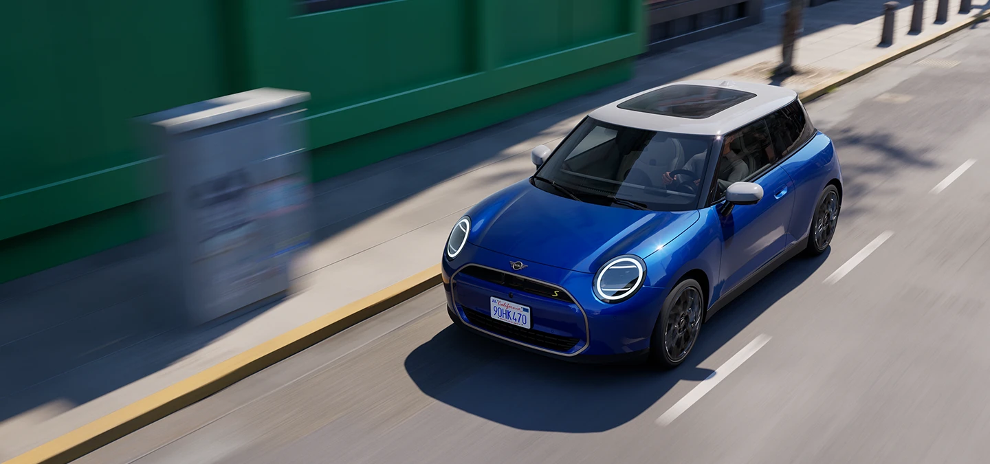 Pohľad zhora na čisto elektrické MINI Cooper vo farbe Blazing Blue so strechou White, počas jazdy na ceste. 