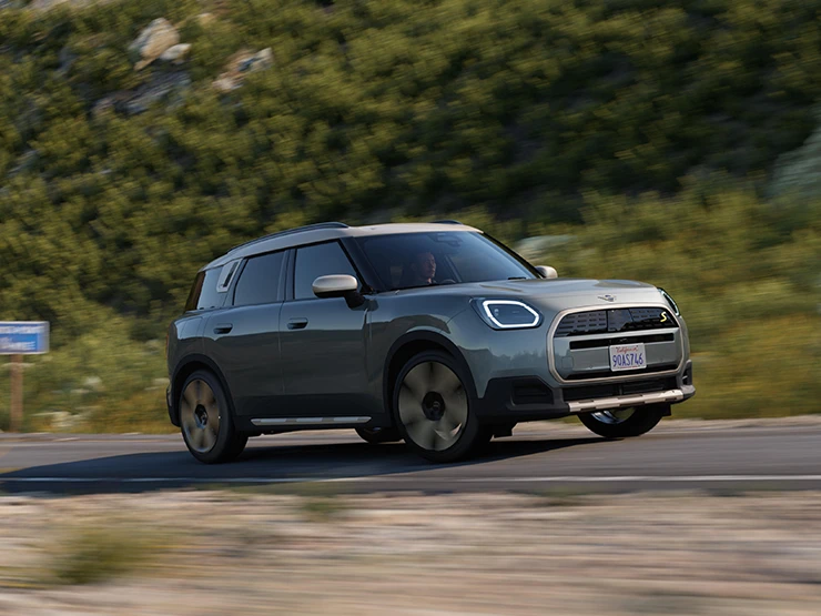Bočný jazdný pohľad na čisto elektrické MINI Countryman vo farbe Smokey Green so strechou Vibrant Silver, v teréne.