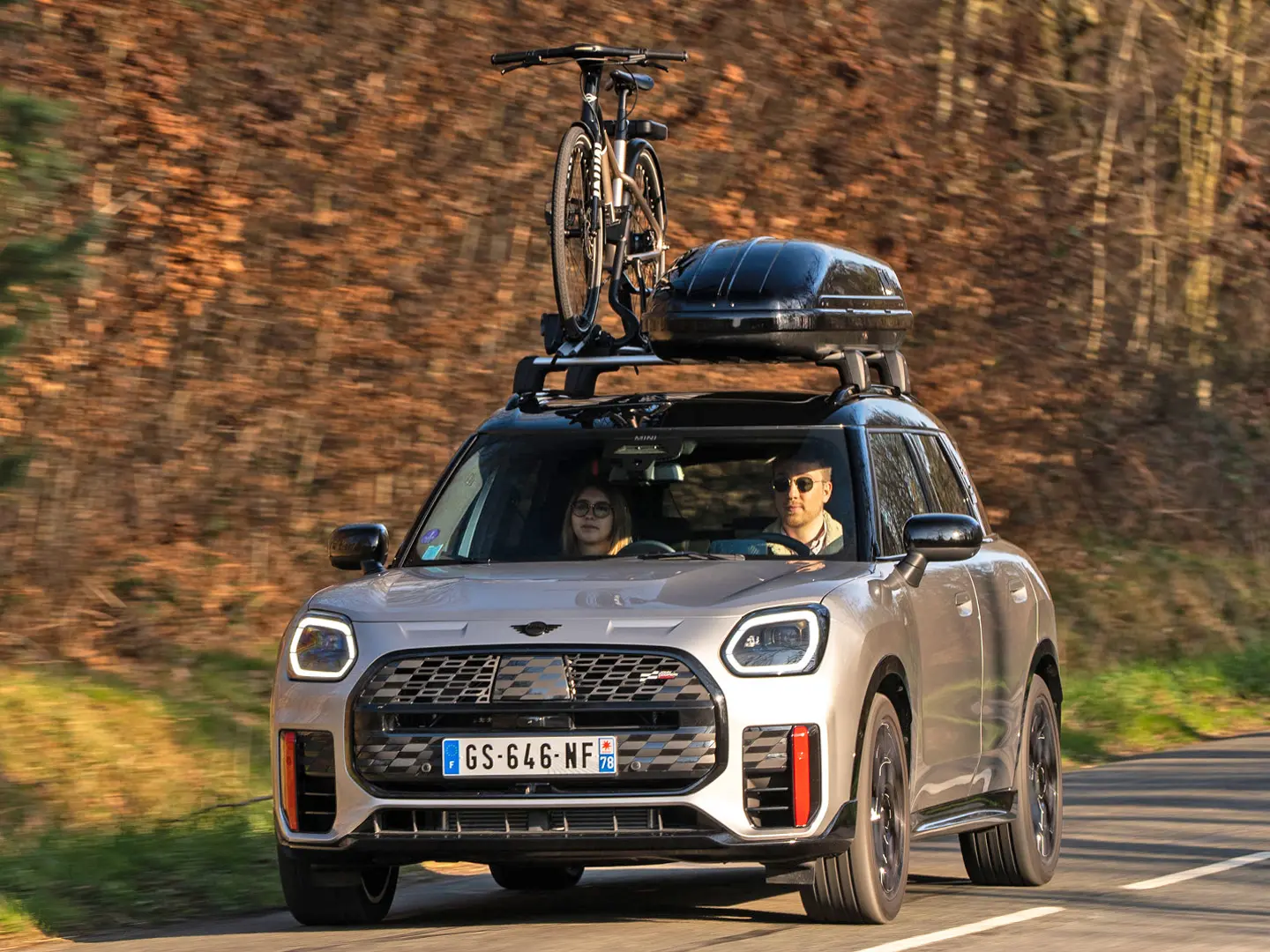 MINI Countryman Cooper v striebornej farbe na horskej ceste so strešným nosičom MINI, strešným boxom MINI, strešným nosičom na bicykle MINI s jedným nainštalovaným bicyklom a s aerodynamickou lištou MINI John Cooper Works – pohľad spredu. 
