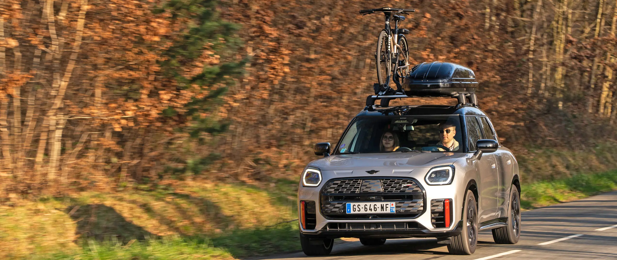 MINI Countryman Cooper v striebornej farbe na horskej ceste so strešným nosičom MINI, strešným boxom MINI, strešným nosičom na bicykle MINI s jedným nainštalovaným bicyklom a s aerodynamickou lištou MINI John Cooper Works – pohľad spredu. 