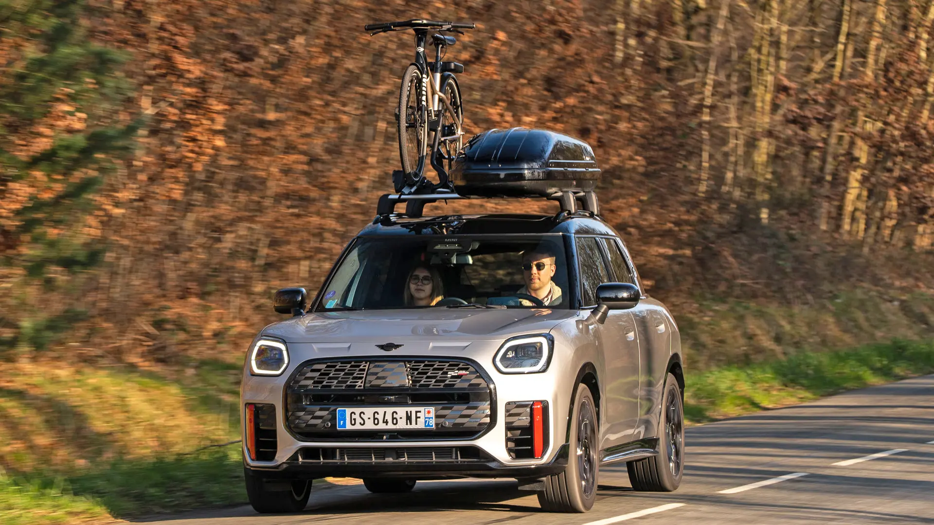 MINI Countryman Cooper v striebornej farbe na horskej ceste so strešným nosičom MINI, strešným boxom MINI, strešným nosičom na bicykle MINI s jedným nainštalovaným bicyklom a s aerodynamickou lištou MINI John Cooper Works – pohľad spredu. 