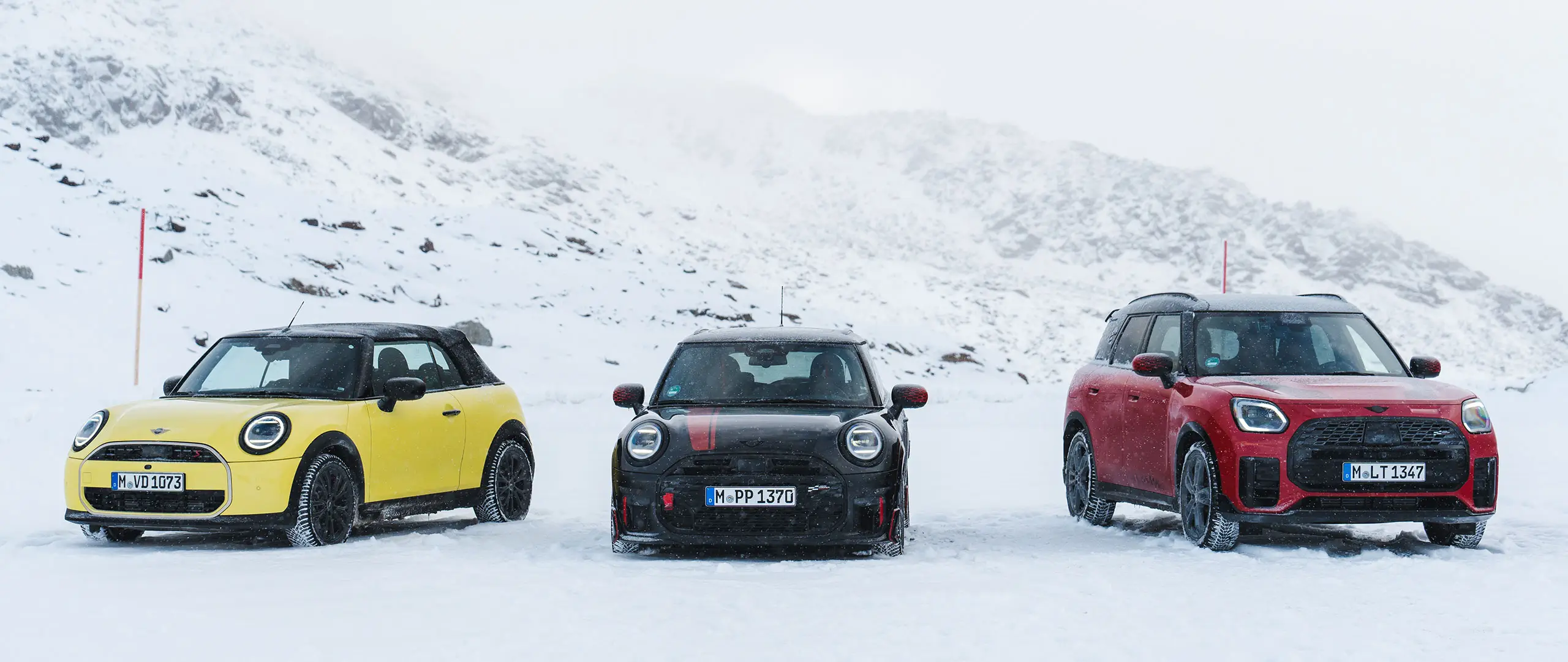MINI Cooper Cabrio vo farbe Sunny Side Yellow, MINI John Cooper Works vo farbe Midnight Black II a MINI Countryman Cooper vo farbe Chili Red – stojace na zasneženej horskej ceste, pohľad spredu.